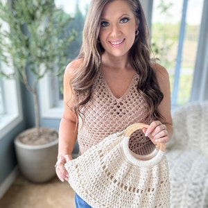 May include: A woman is holding a crocheted tote bag with a wooden handle. The bag is cream colored and has a delicate open stitch pattern. The woman is wearing a brown crochet top and blue jeans.