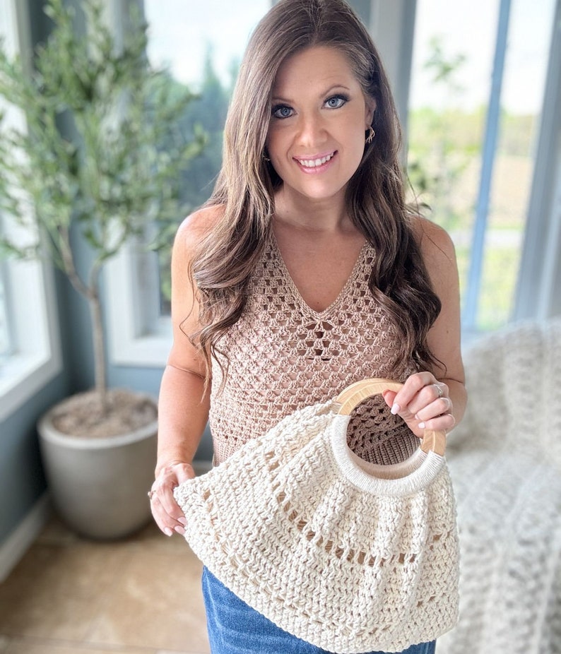 May include: A woman is holding a crocheted tote bag with a wooden handle. The bag is cream colored and has a delicate open stitch pattern. The woman is wearing a brown crochet top and blue jeans.
