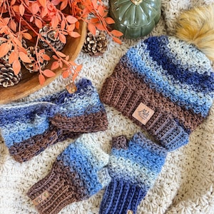 May include: A crochet set in shades of blue and brown, including a beanie with a faux fur pom, a headband, and fingerless gloves. Each item has a small leather tag. The set is displayed on a cream-colored, crocheted blanket.