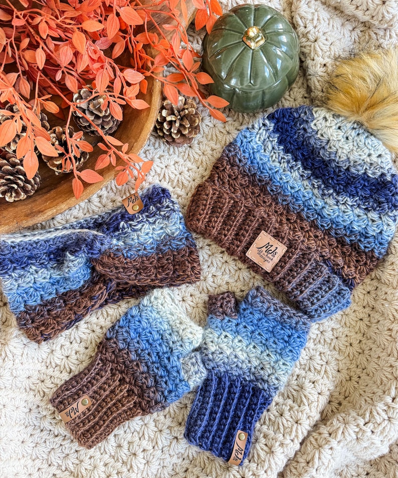 May include: A crochet set in shades of blue and brown, including a beanie with a faux fur pom, a headband, and fingerless gloves. Each item has a small leather tag. The set is displayed on a cream-colored, crocheted blanket.