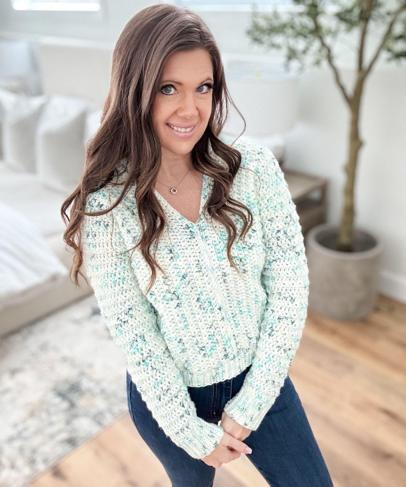 May include: A white crocheted sweater with a green and blue speckled pattern. The sweater has a v-neck and long sleeves.