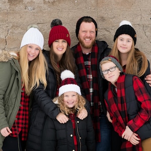 May include: A family of six wearing red and black plaid shirts and matching knit hats with pom-poms. The hats are in various colors, including white, red, and black. The family is standing in front of a concrete wall.