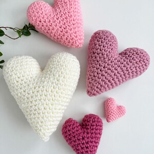 Pu&ograve; includere: Cinque cuori all'uncinetto di diverse dimensioni e tonalit&agrave; di rosa e bianco. I cuori sono disposti su una superficie bianca.