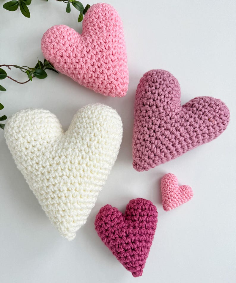 Pu&ograve; includere: Cinque cuori all'uncinetto di diverse dimensioni e tonalit&agrave; di rosa e bianco. I cuori sono disposti su una superficie bianca.
