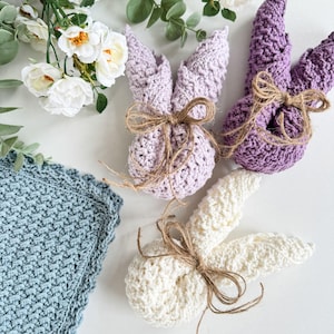 May include: Three crocheted bunny rabbits in purple, white, and light blue. Each bunny has a brown twine bow tied around its neck.