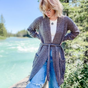 Op de afbeelding: Een lange, grijze cardigan met een strikceintuur en zakken. De cardigan is gemaakt van een zacht, getextureerd garen en heeft een ontspannen pasvorm.