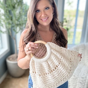 May include: A crocheted tote bag with a wooden handle. The bag is cream colored and has a delicate openwork design. The bag is being held by a person wearing a brown crochet top and blue jeans.