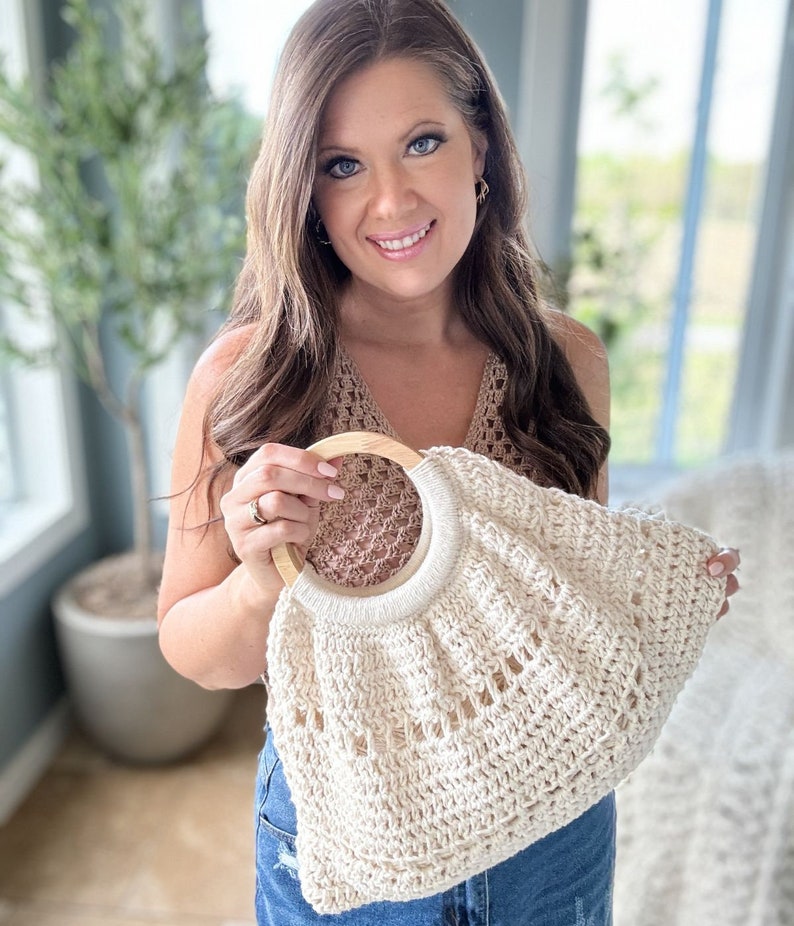 May include: A crocheted tote bag with a wooden handle. The bag is cream colored and has a delicate openwork design. The bag is being held by a person wearing a brown crochet top and blue jeans.