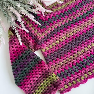 Op de afbeelding: Een handgemaakte gehaakte top in een granny square patroon. De top heeft een kleurengradient van roze, groen en paarse tinten. De mouwen zijn lang en de halslijn is geschulpt. De top wordt gepresenteerd op een wit oppervlak met bloemaccenten.