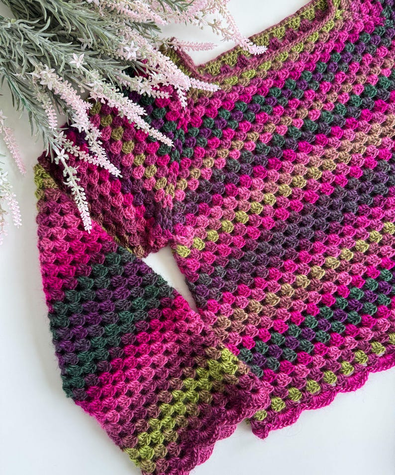 Op de afbeelding: Een handgemaakte gehaakte top in een granny square patroon. De top heeft een kleurengradient van roze, groen en paarse tinten. De mouwen zijn lang en de halslijn is geschulpt. De top wordt gepresenteerd op een wit oppervlak met bloemaccenten.