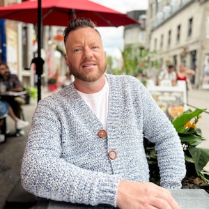 May include: A light blue and white speckled knit cardigan with two brown buttons. The cardigan is worn over a white t-shirt. The image is taken outdoors, with a red umbrella and a city street in the background.