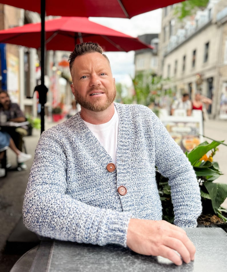 May include: A light blue and white speckled knit cardigan with two brown buttons. The cardigan is worn over a white t-shirt. The image is taken outdoors, with a red umbrella and a city street in the background.