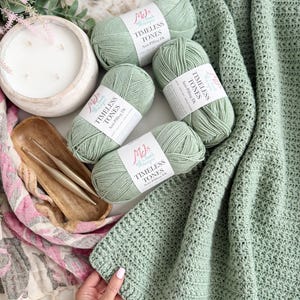 Puede incluir: Cuatro ovillos de lana verde salvia con etiquetas blancas que dicen MJs Off The Hook Designs Timeless Tones Anti-Pilling Dk, dos ganchillos dorados en un plato de madera y una manta de ganchillo de color verde salvia texturizada.