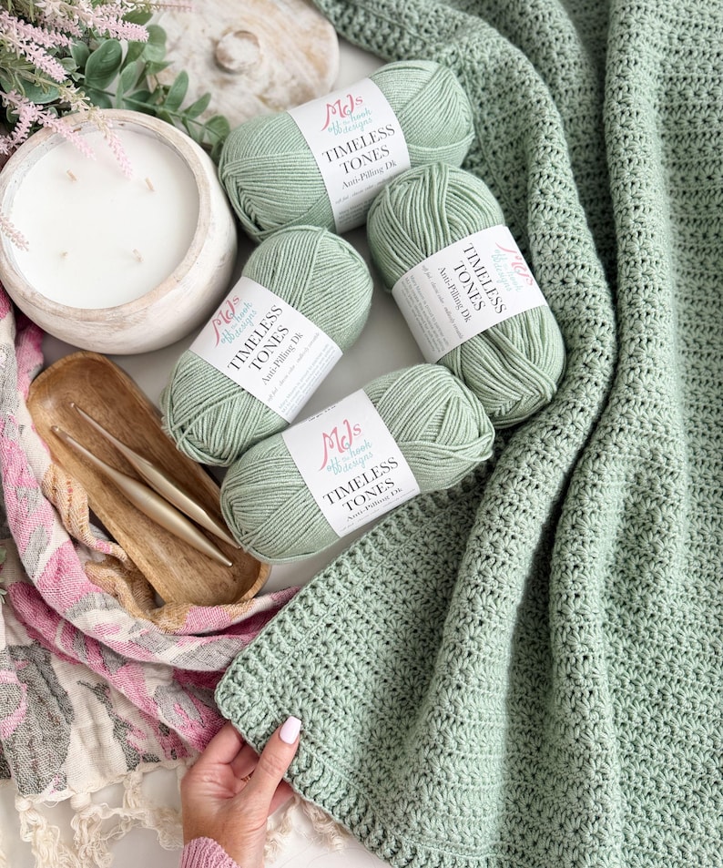 Puede incluir: Cuatro ovillos de lana verde salvia con etiquetas blancas que dicen MJs Off The Hook Designs Timeless Tones Anti-Pilling Dk, dos ganchillos dorados en un plato de madera y una manta de ganchillo de color verde salvia texturizada.