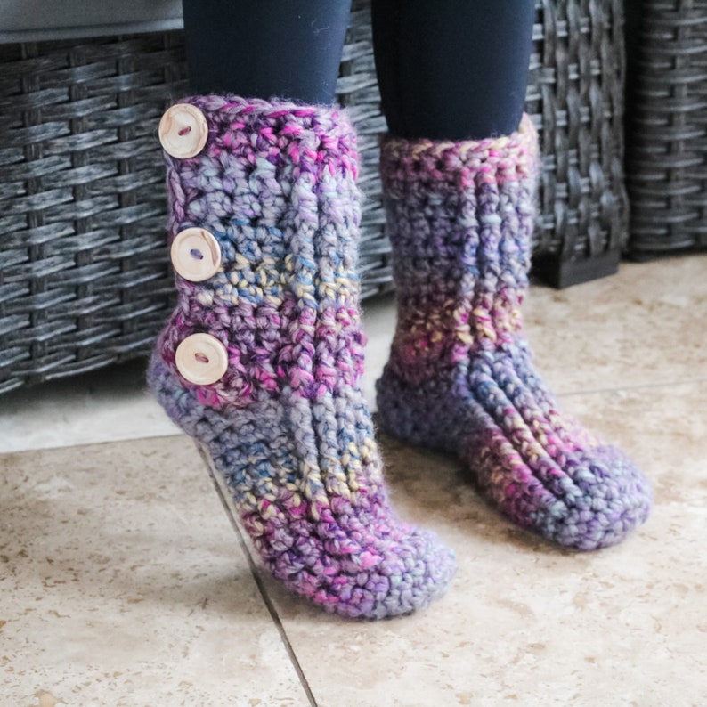 May include: Purple and blue crocheted slipper socks with a button detail. The socks have a textured knit pattern and are made with a soft yarn.