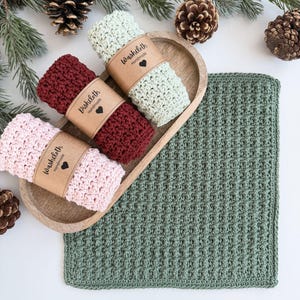May include: A wooden tray holds three rolled washcloths in pink, burgundy, and light green, each with a paper band that reads "Washcloth Handmade" and a heart. A larger green washcloth lies beside the tray, with pinecones and greenery in the background.