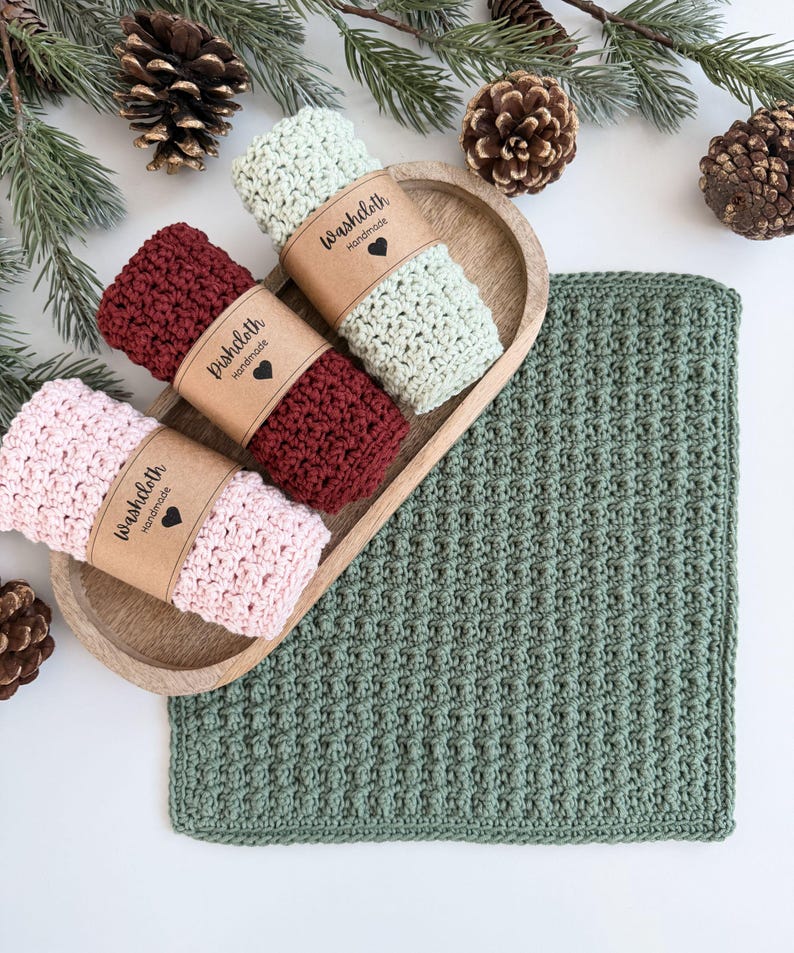 May include: A wooden tray holds three rolled washcloths in pink, burgundy, and light green, each with a paper band that reads "Washcloth Handmade" and a heart. A larger green washcloth lies beside the tray, with pinecones and greenery in the background.