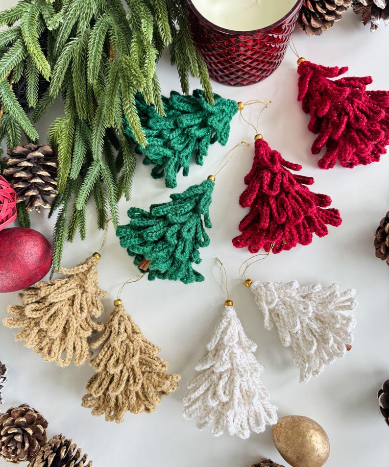 K&ouml;nnte beinhalten: Handgefertigte Weihnachtsbaumschmuck aus H&auml;kelarbeit in Gr&uuml;n, Rot, Beige und Wei&szlig;. Jeder Baum hat eine goldfarbene Spitze und eine Schlaufe zum Aufh&auml;ngen. Die Ornamente sind mit Tannenzapfen und Gr&uuml;n arrangiert.