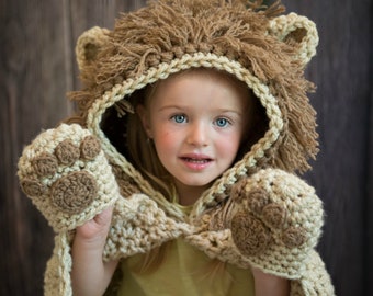 May include: A child wearing a crocheted lion costume with a brown and cream color scheme. The costume features a hood with lion ears and a mane, as well as paw-shaped mittens.
