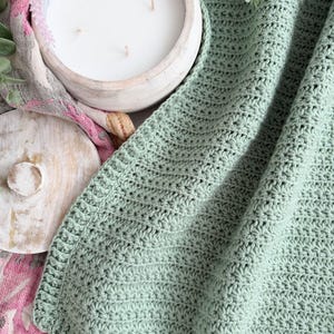 Puede incluir: Una manta de ganchillo color verde salvia con un patr&oacute;n de puntos de estrella texturizados y un borde acanalado. La manta est&aacute; colocada junto a una vela blanca de tres mechas en un cuenco de madera redondo y una tela con estampado floral.