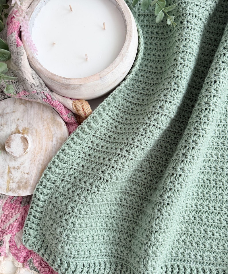 Puede incluir: Una manta de ganchillo color verde salvia con un patr&oacute;n de puntos de estrella texturizados y un borde acanalado. La manta est&aacute; colocada junto a una vela blanca de tres mechas en un cuenco de madera redondo y una tela con estampado floral.