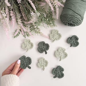Può includere: Trifogli fatti a mano all'uncinetto in varie tonalità di verde, disposti su una superficie bianca. Sono presenti anche un gomitolo di filo verde e rametti di fiori rosa e verdi. Un trifoglio è tenuto da una persona.