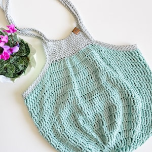 May include: A crocheted tote bag with a light green and gray color scheme. The bag has a wide, flat base and two long straps.