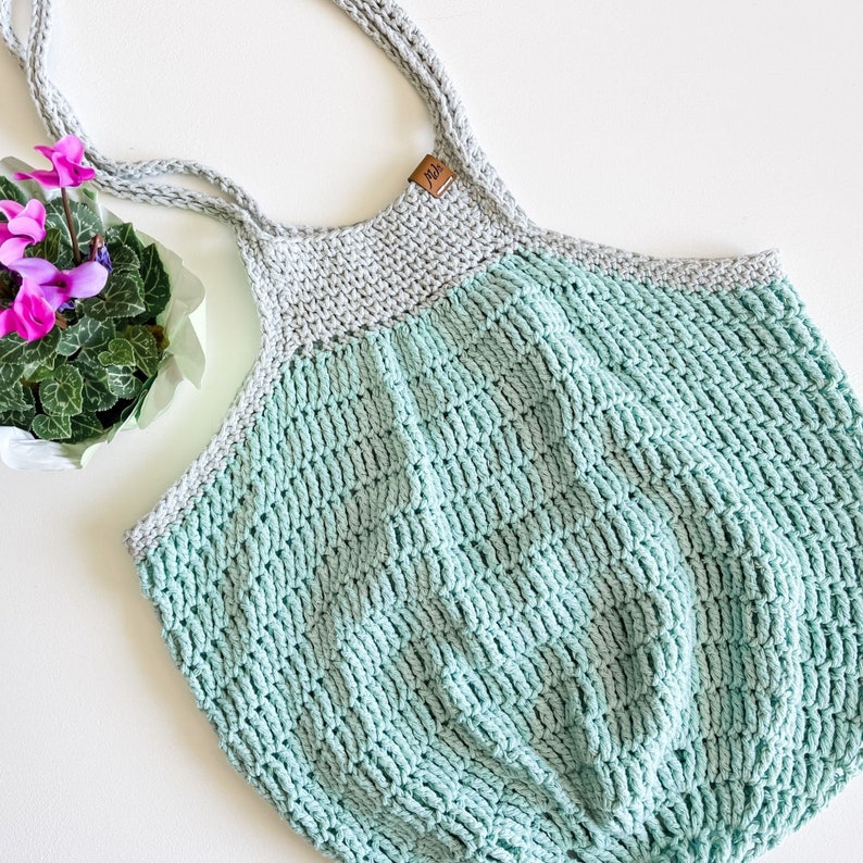 May include: A crocheted tote bag with a light green and gray color scheme. The bag has a wide, flat base and two long straps.