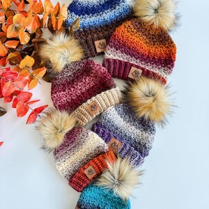 May include: A collection of handmade knit beanies in various colors, including blue, orange, and burgundy. Each beanie features a faux fur pom-pom and a small, rectangular tag. The beanies are arranged on a white surface.