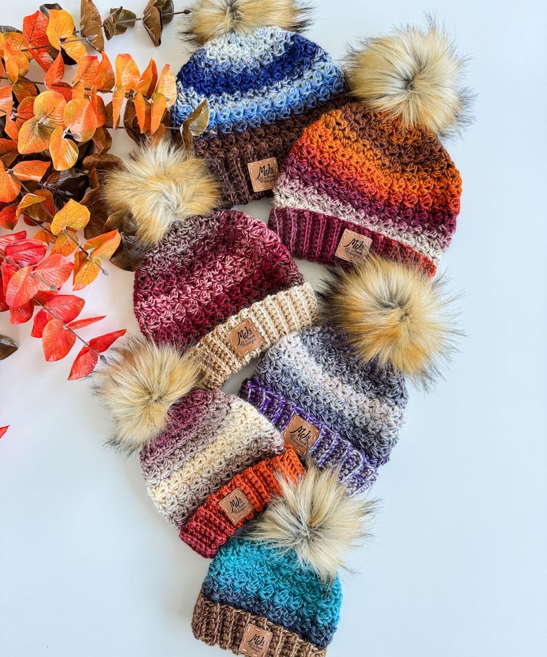 May include: A collection of handmade knit beanies in various colors, including blue, orange, and burgundy. Each beanie features a faux fur pom-pom and a small, rectangular tag. The beanies are arranged on a white surface.