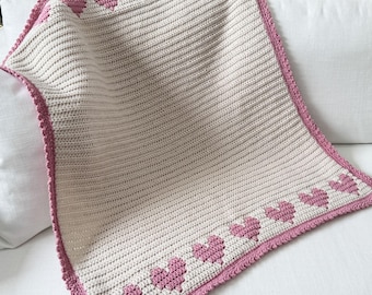 May include: A crocheted baby blanket in cream with a pink border and pink hearts along the bottom edge.