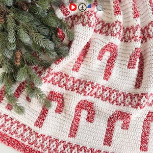 K&ouml;nnte beinhalten: Ein wei&szlig;-roter geh&auml;kelter Weihnachtsbaumrock mit einem Zuckerstangenmuster. Der Rock hat einen roten Rand und ist &uuml;ber einer Oberfl&auml;che mit einem Zweig aus immergr&uuml;nen Pflanzen und Tannenzapfen drapiert.