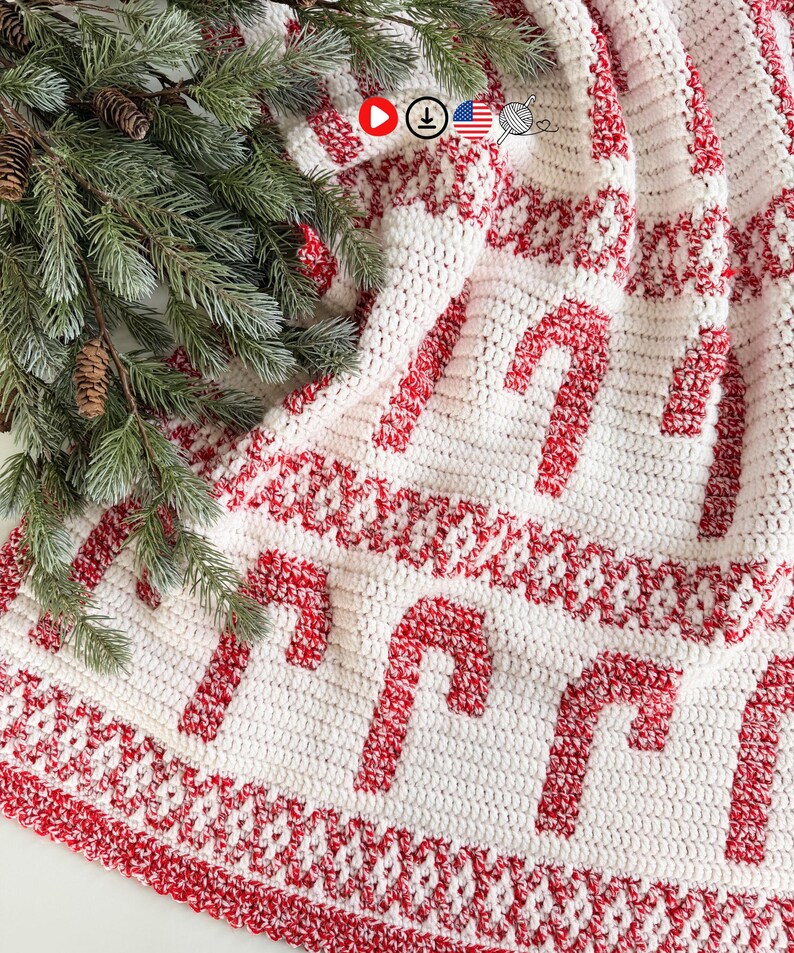 K&ouml;nnte beinhalten: Ein wei&szlig;-roter geh&auml;kelter Weihnachtsbaumrock mit einem Zuckerstangenmuster. Der Rock hat einen roten Rand und ist &uuml;ber einer Oberfl&auml;che mit einem Zweig aus immergr&uuml;nen Pflanzen und Tannenzapfen drapiert.