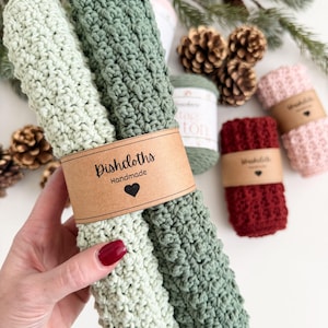 May include: Handmade dishcloths in shades of green, red, and pink, with a textured crochet design. The dishcloths are bundled with a brown paper band that reads "Dishcloths Handmade" with a heart symbol. Pinecones and greenery are in the background.