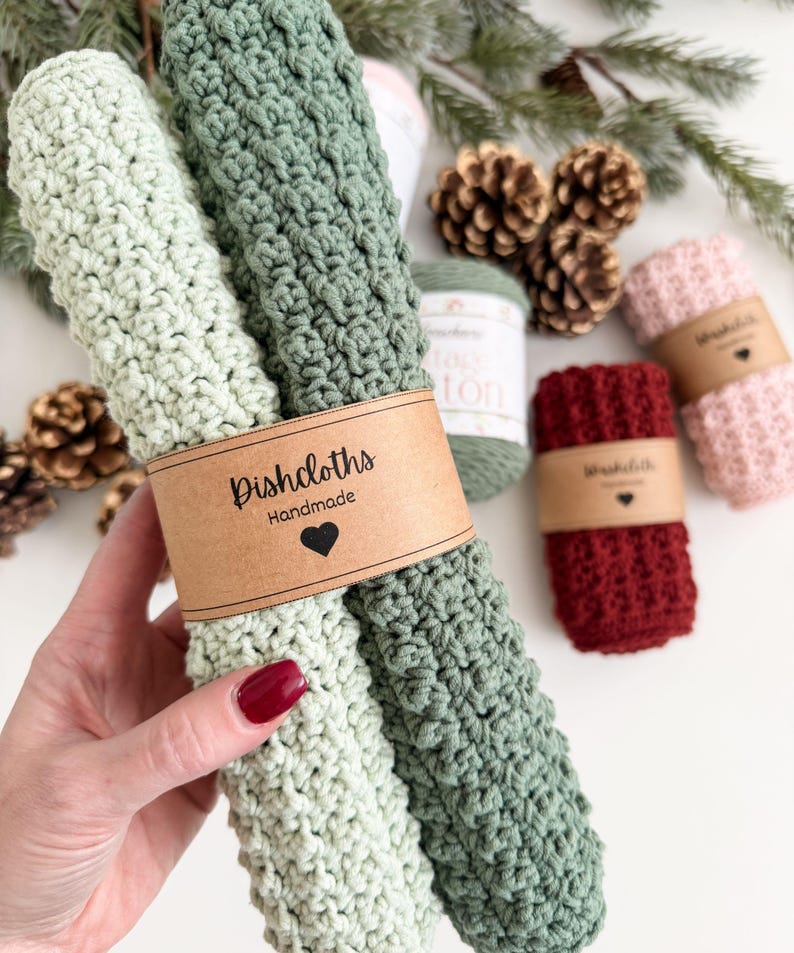 May include: Handmade dishcloths in shades of green, red, and pink, with a textured crochet design. The dishcloths are bundled with a brown paper band that reads "Dishcloths Handmade" with a heart symbol. Pinecones and greenery are in the background.