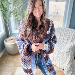 K&ouml;nnte beinhalten: Eine Frau tr&auml;gt eine braune, blaue und wei&szlig;e geh&auml;kelte Strickjacke mit langem &Auml;rmel. Sie tr&auml;gt eine blaue Jeans und hat lange braune Haare.