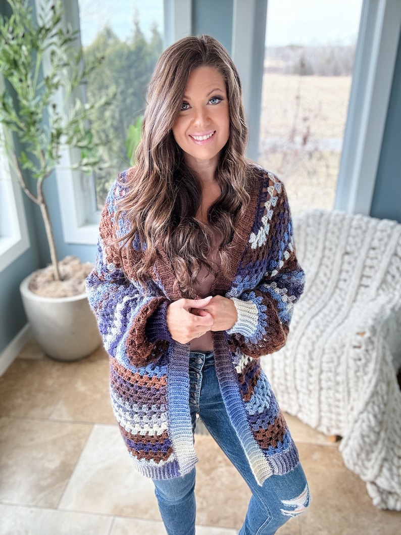 K&ouml;nnte beinhalten: Eine Frau tr&auml;gt eine braune, blaue und wei&szlig;e geh&auml;kelte Strickjacke mit langem &Auml;rmel. Sie tr&auml;gt eine blaue Jeans und hat lange braune Haare.