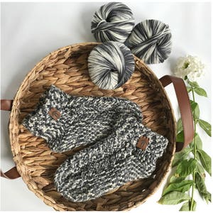 May include: Two pairs of black and white knitted socks with a brown leather tag. The socks are in a woven basket with three balls of yarn.