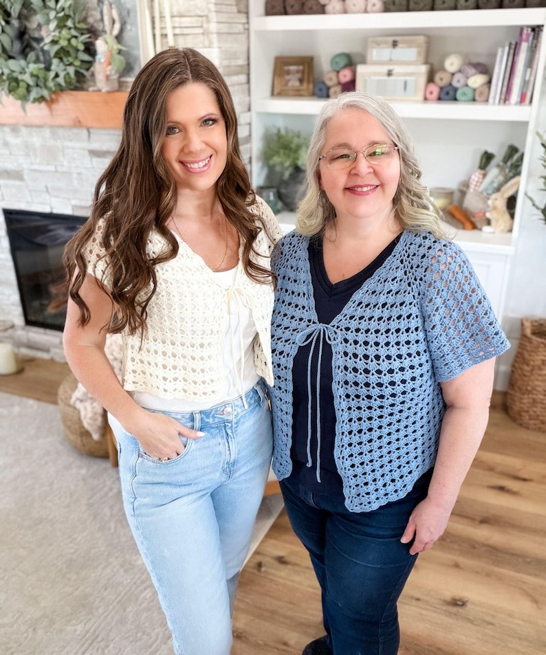 May include: Two women wearing handmade crochet cardigans. One cardigan is cream-colored, the other is light blue. Both cardigans have a tie closure. They are standing in front of a fireplace and a white bookshelf.