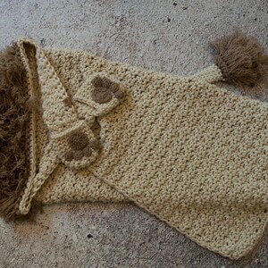 May include: A crocheted lion hooded blanket in beige with brown paw appliques. The blanket has a long tail with a brown tassel.