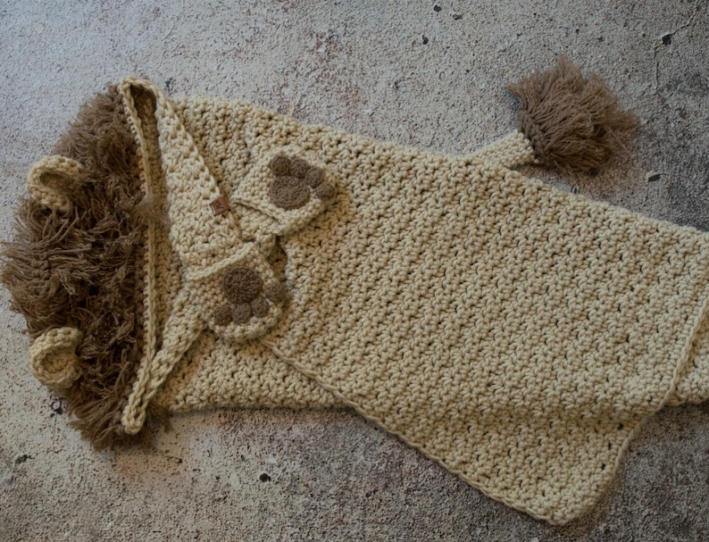 May include: A crocheted lion hooded blanket in beige with brown paw appliques. The blanket has a long tail with a brown tassel.