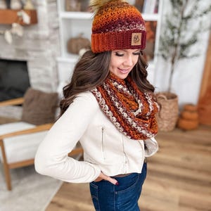 May include: A knitted hat and infinity scarf set in shades of orange, burgundy, and cream. The hat features a faux fur pom-pom and a small leather patch. The scarf is a looped design, and the set appears to be handmade.