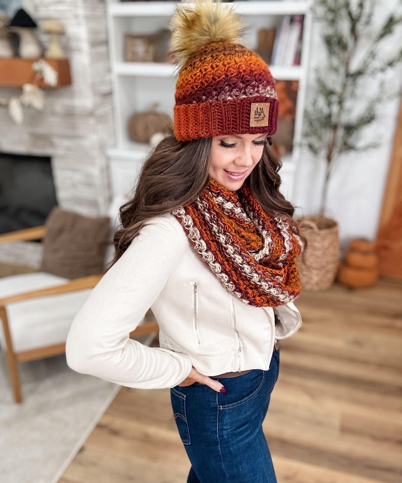 May include: A knitted hat and infinity scarf set in shades of orange, burgundy, and cream. The hat features a faux fur pom-pom and a small leather patch. The scarf is a looped design, and the set appears to be handmade.
