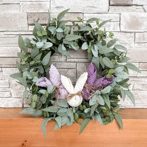 May include: A green eucalyptus wreath with two crocheted bunny decorations. The bunnies are purple and white and have a rustic twine bow.