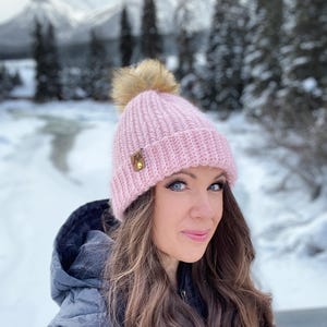 Peut inclure: Un bonnet en crochet rose avec un pompon en fausse fourrure. Le bonnet a une petite étiquette en cuir marron avec le mot "Made" dessus. Le bonnet est porté par une personne aux longs cheveux bruns, debout devant un fond enneigé.