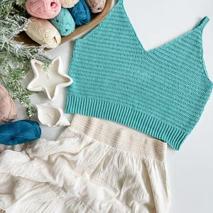 Pu&ograve; includere: Un top all'uncinetto turchese con scollo a V e una gonna color crema con orlo a balze. L'immagine presenta anche una ciotola di legno con vari colori di filato, tra cui rosa, verde acqua e beige, e candele decorative.