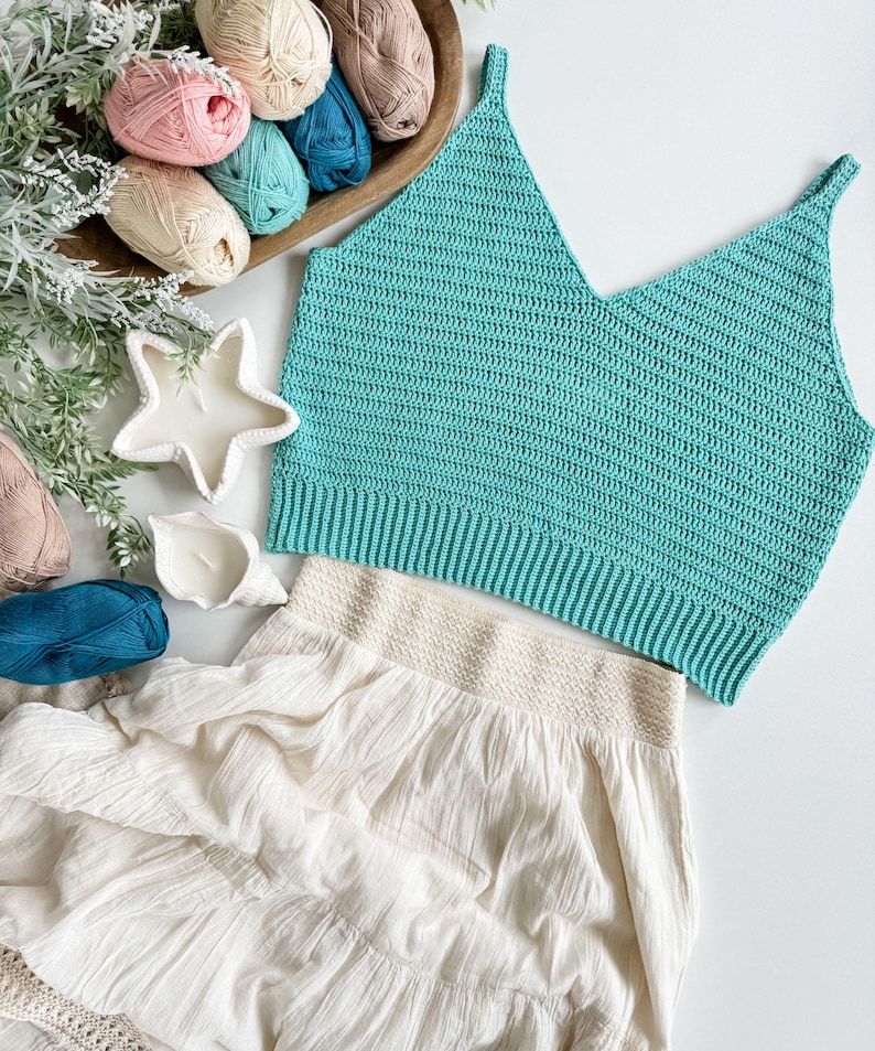 Pu&ograve; includere: Un top all'uncinetto turchese con scollo a V e una gonna color crema con orlo a balze. L'immagine presenta anche una ciotola di legno con vari colori di filato, tra cui rosa, verde acqua e beige, e candele decorative.