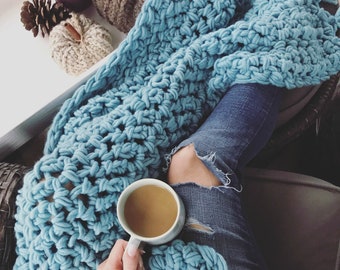 May include: A person is sitting on a couch with a blue crocheted blanket and a cup of coffee. The blanket is made with chunky yarn and has a loose, open weave. The person is wearing blue jeans with rips in the knees.