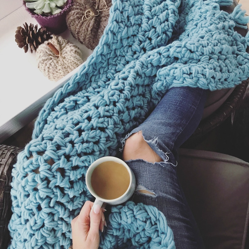 Pu&ograve; includere: Una persona &egrave; seduta su un divano con una coperta all'uncinetto blu e una tazza di caff&egrave;. La coperta &egrave; realizzata con filato spesso e ha una trama sciolta e aperta. La persona indossa jeans blu con strappi alle ginocchia.