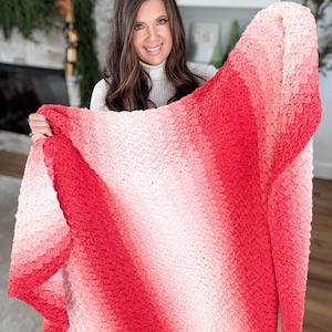 May include: A handmade, crochet blanket with a gradient of colors from light pink to deep red. The blanket has a textured stitch pattern and is displayed against a neutral background. The blanket is a cozy home decor item.